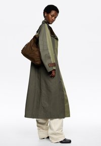 Oliwkowy długi płaszcz z brązowymi akcentami, z szerokim kołnierzem i luźnym krojem. Połączony z beżowymi spodniami i brązową torbą na ramię.