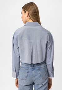 Lys blå, cropped skjorte med knapper og lange ærmer, en rygfald og opklippede ærmer. Parret med højhalsede denim jeans.