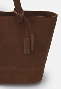 Brun mockasin tote-väska med dubbel läderfransdetalj hängande från en rem, med sydda kanter längs de övre och nedre kanterna.