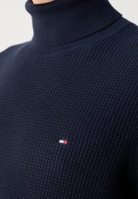 Gros plan sur un pull à col roulé texturé bleu marine avec un petit logo rectangulaire rouge, blanc et bleu sur la poitrine.