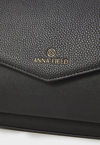 Pochette synthétique noire texturée avec une section inférieure lisse, arborant un logo doré "ANNA FIELD" à l'avant. Design élégant avec un rabat triangulaire.