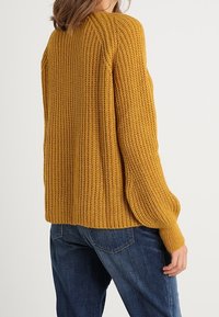 Senfgelbes gestricktes Sweater mit geripptem Muster, lockerer Passform und überschnittenen Schultern. Kombiniert mit blauen Jeans.