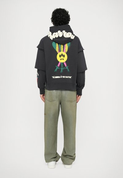 Felpa nera con grafica colorata che presenta un sorriso giallo e foglie. Abbinata a jeans larghi verde oliva e sneaker bianche.
