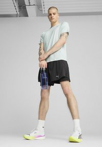 Lys turkis atletisk skjorte, sorte shorts, hvide og neongrønt gradient sneakers, holder en klar vandflaske, står på en neutral baggrund.