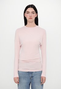 Pale pink long-sleeve top features a round neckline, fitted silhouette, and ruched detailing. Paired with light blue denim jeans.