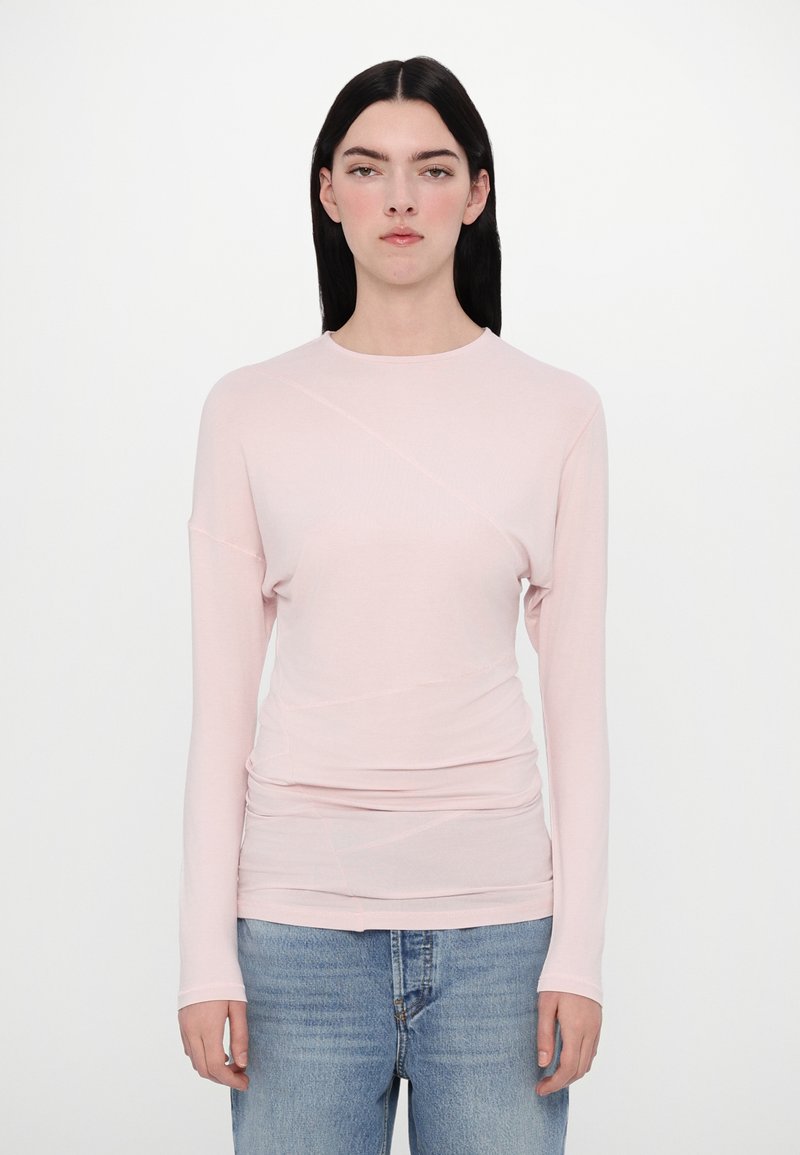 Pale pink long-sleeve top features a round neckline, fitted silhouette, and ruched detailing. Paired with light blue denim jeans.