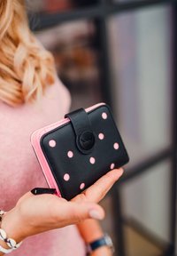 Black and pink wallet featuring a polka dot pattern, leather material, a flap closure, and a circular logo accent. Held in a hand.