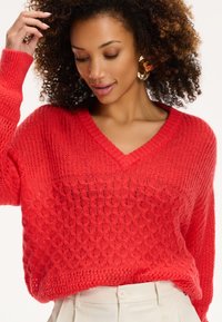 Maglione rosso lavorato a maglia con scollatura a V, modello a fantasia testurizzata e vestibilità ampia. Presenta polsini a coste e una silhouette morbida. Indossato con pantaloni color crema.