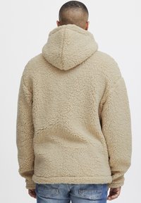 Beige fleece hettegenser med en teksturert overflate, stor hette og en avslappet passform, kombinert med blå jeans. Materialet ser mykt og koselig ut.