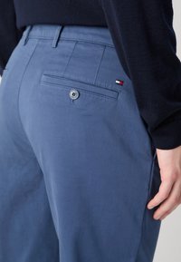 Pantaloni blu realizzati in tessuto con una texture liscia, caratterizzati da una tasca posteriore con bottone e un piccolo logo in rosso, bianco e blu.