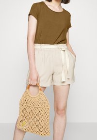 Hnědý top s krátkým rukávem, béžové šortky s elastickým pasem a stojícím uzlem. Drží světle hnědou tkanou tašku s kulatým dřevěným držadlem a diamantovým vzorem.