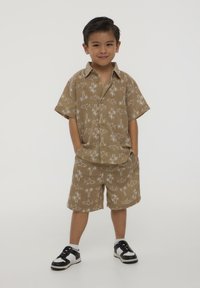 Una maglietta corta beige con maniche corte e pantaloni corti abbinati con un motivo di pesci bianchi e palme, abbinati a scarpe da ginnastica bianche e nere, indossata da un bambino.