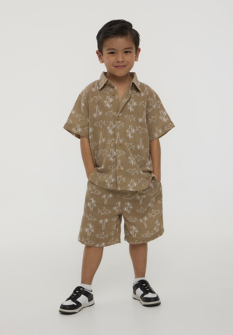 Una maglietta corta beige con maniche corte e pantaloni corti abbinati con un motivo di pesci bianchi e palme, abbinati a scarpe da ginnastica bianche e nere, indossata da un bambino.