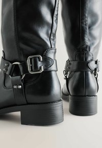 Mustadest nahast põlvini saapad, millel on matt viimistlus, hõbedase tooni raudtee detailid ja veidi tekstureeritud ploki konts.