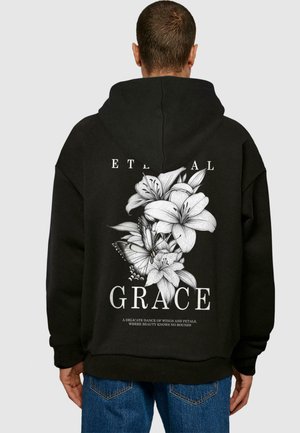 Sort hættetrøje med et hvidt blomstermotiv med liljer og sommerfugle på ryggen, ledsaget af teksten "ETERNAL GRACE."