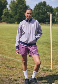 Nike siva bomber jakna s plavim ovratnikom, ljubičaste hlačice s leopard printom, bijele čarape i crne cipele, stoji na travnatoj površini.