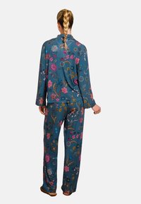 Blaue Stoff-Pyjama-Set mit floraler Stickerei in Pink, Lila und Gelb. Lange Ärmel und lockere Hosen, getragen mit braunen Schuhen.