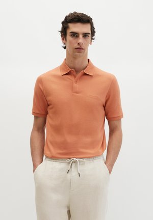 Junger Mann trägt ein kurzärmeliges Polohemd in Lachsfarbe und hellbeige Hose mit Kordelzug, steht mit den Händen in den Taschen vor schlichten Hintergrund.