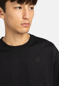 Zwarte katoenen t-shirt met een ronde hals, korte mouwen en een geborduurd logo-accent op het linkerborstgedeelte. Gladde textuur.
