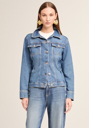 Woman wearing fitted blue denim jacket and matching jeans, accessorized with large gold flower-shaped earrings, standing against a plain background.