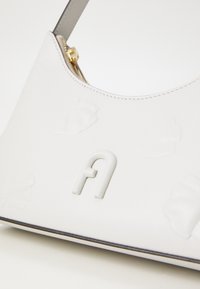 Witte leren handtas met een gebogen vorm, voorzien van reliëf bloemenpatronen en een verhoogd logo in het midden. Grijze schouderband en gouden hardware.