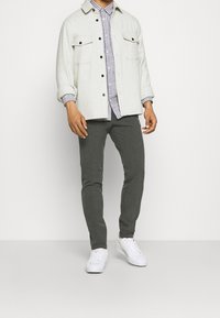 Grå byxor som matchas med en vit hållbar jacka med två bröstfickor och svarta knappar över en randig skjorta. Vita sneakers fulländar looken.