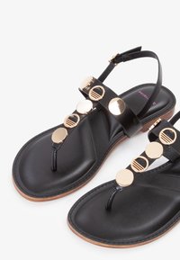 Black leather sandals feature a thong design with gold circular accents along the straps and a flat sole with a natural-colored edge.
