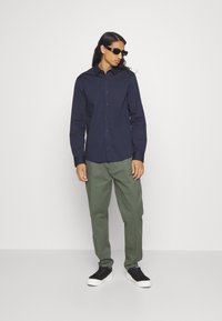 Camicia button-up navy in cotone, abbinata a pantaloni verde oliva e sneakers neri. Il modello indossa occhiali da sole neri, in piedi su uno sfondo neutro.
