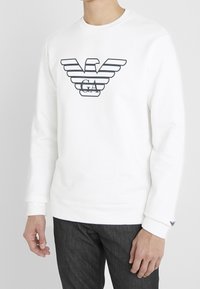 White sweatshirt with a crew neck, featuring a black striped eagle logo and “GA” text. Soft texture and ribbed cuffs, paired with gray jeans.