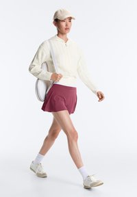 Femme marchant portant un pull en maille crème, une jupe bordeaux, des baskets blanches, des chaussettes blanches, une casquette beige et un sac rond blanc sur l’épaule.