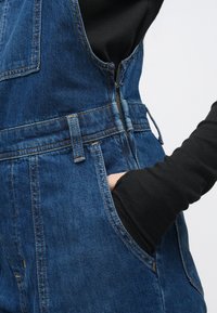 Τζιν σαλοπέτα σκούρου μπλε χρώματος με εφαρμοστή σχεδίασ�η, διαθέτει φερμουάρ, τσέπες και αντίθετη πορτοκαλί ραφή. Μαύρο μπλουζάκι με μακριά μανίκια από κάτω.