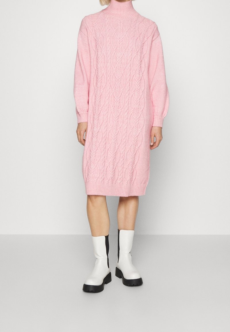 Pink knitted sweater dress with cable pattern, high neckline, and ribbed cuffs, paired with white and black ankle boots.