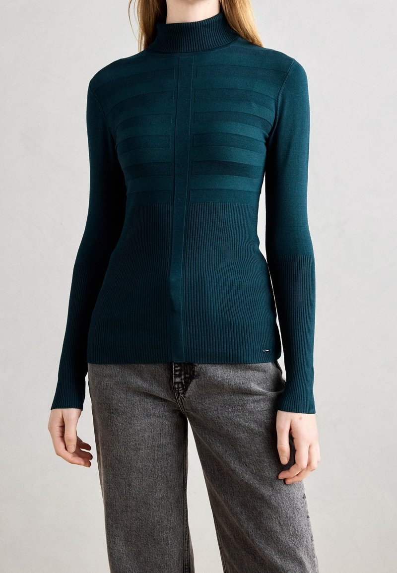 Femme portant un pull à col roulé côtelé à manches longues de couleur sarcelle et un jean gris taille haute, debout devant un fond clair uni.