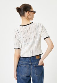 Weißes gehäkeltes Oberteil mit schwarzem Besatz, kurzen Ärmeln und cropped Design, kombiniert mit hoch taillierten blauen Jeans mit klassischen Taschen.
