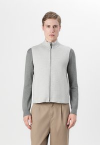 Pelēks zip-up veste ar gaiši pelēku priekšpusi un tumšākām pelēkām piedurknēm, ar augstu apkakli un ribota tipa aprocēm, apvienota ar gaiši brūnām biksēm.