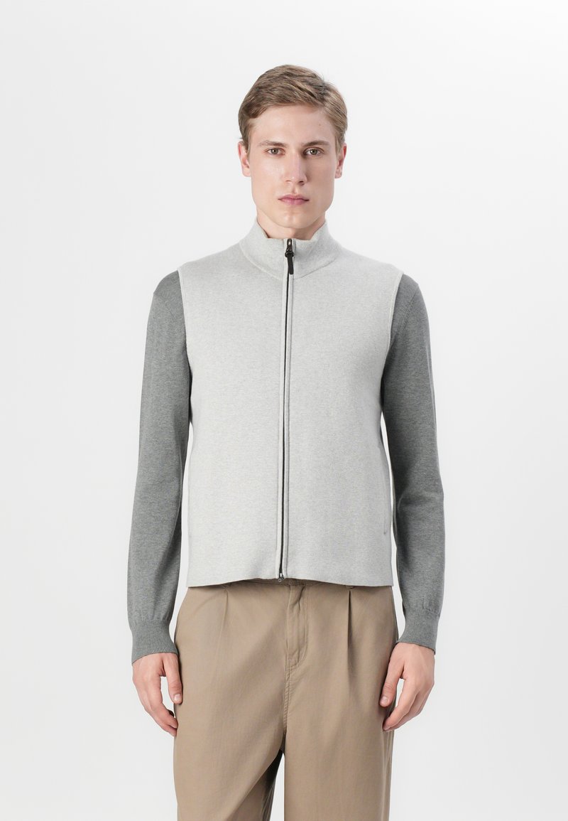 Pelēks zip-up veste ar gaiši pelēku priekšpusi un tumšākām pelēkām piedurknēm, ar augstu apkakli un ribota tipa aprocēm, apvienota ar gaiši brūnām biksēm.
