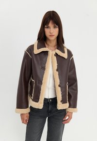 Brun kunstlærjakke med beige pelsfor, stor krage og knappelukking. Har frontlommer og kontrastsøm.