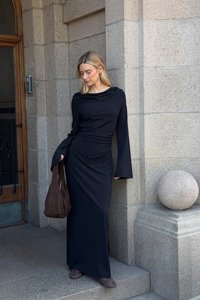 Svart långärmad klänning med draperad halsringning och åtsydd midja. Modellen håller en brun tote-väska, bär bruna skor, mot en stenmur.