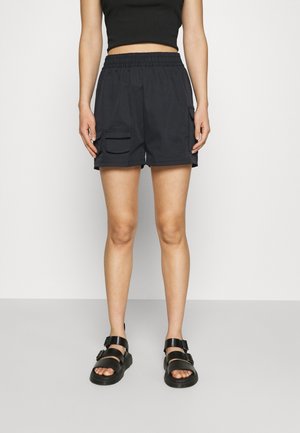 Person iført sorte elastiske shorts med sidelommer, en sort cropped top og sorte åbne sandaler med spænder, stående.