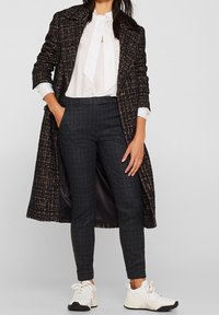 Vrouw in een gestructureerde lange jas, witte blouse met strik, donkere geruite broek en witte sneakers, staand met één hand in de zak.