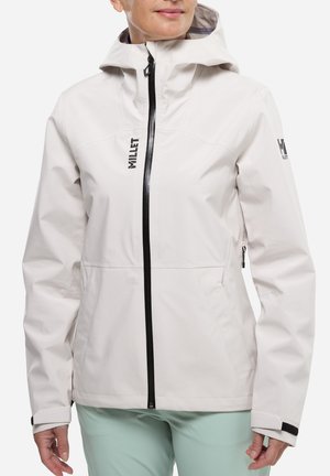 Veste à capuche gris clair avec fermeture éclair noire à l'avant et logo sur la poitrine et la manche, portée par une personne avec un pantalon bleu clair, debout devant un fond blanc.