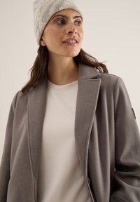 Manteau gris ajusté avec une texture douce, doté d'un col cranté et d'un design ajusté, porté sur un haut clair et un bonnet en tricot crème.