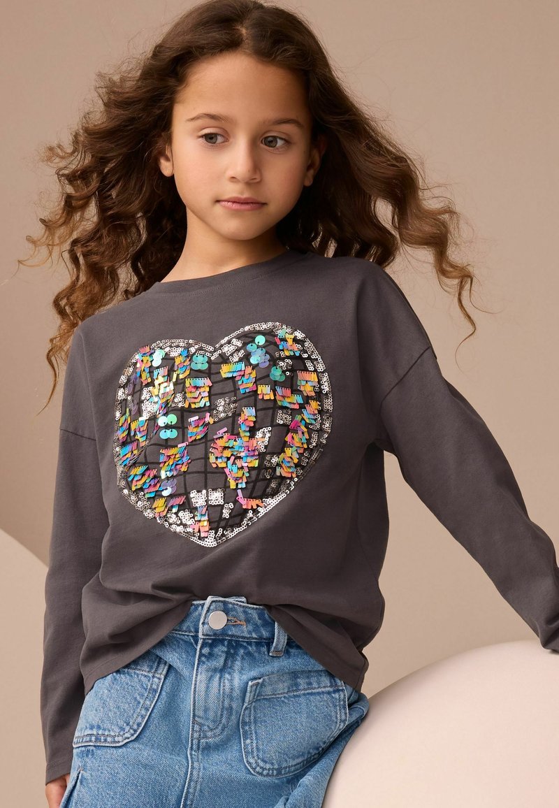 Jeune fille aux longs cheveux bouclés portant un sweat-shirt gris avec un motif cœur coloré en sequins et une jupe en jean, debout devant un fond beige.