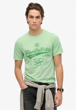 Camiseta de algodón verde claro con mangas cortas y cuello redondo, que presenta gráficos de texto en verde y negro, usada sobre jeans oscuros con un suéter gris atado a la cintura.