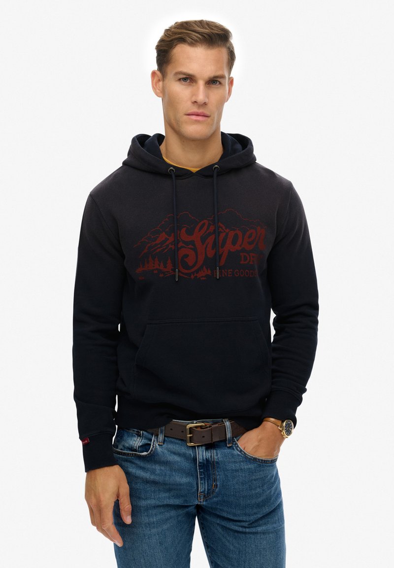 Mørkeblå hættetrøje med snore og en forlommeslomme. Har et rødt bjerggrafik og "Superdry" tekst. Båret sammen med lyseblå jeans.
