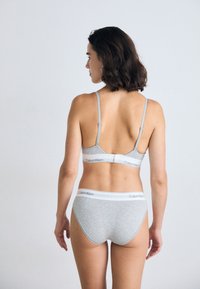 Vrouw die met de rug naar de camera staat in een grijze Calvin Klein beha en bijpassend ondergoed tegen een effen lichte achtergrond.