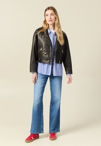 Chaqueta de motocicleta de cuero negro, corta, con cremalleras plateadas, camisa de botones a rayas azul y blanca, jeans azul acampanados y zapatillas rojas.