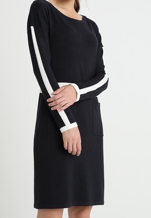 Woman in black knee-length dress with white vertical stripes on sleeves and horizontal stripe at waist, standing with arms crossed.