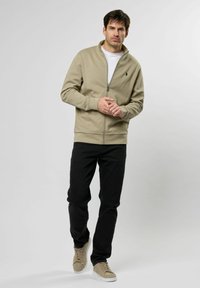 Khaki lynlås hættetrøje med høj krave, ribbede manchetter og et lille logo på brystet, parret med sorte jeans og beige sneakers.