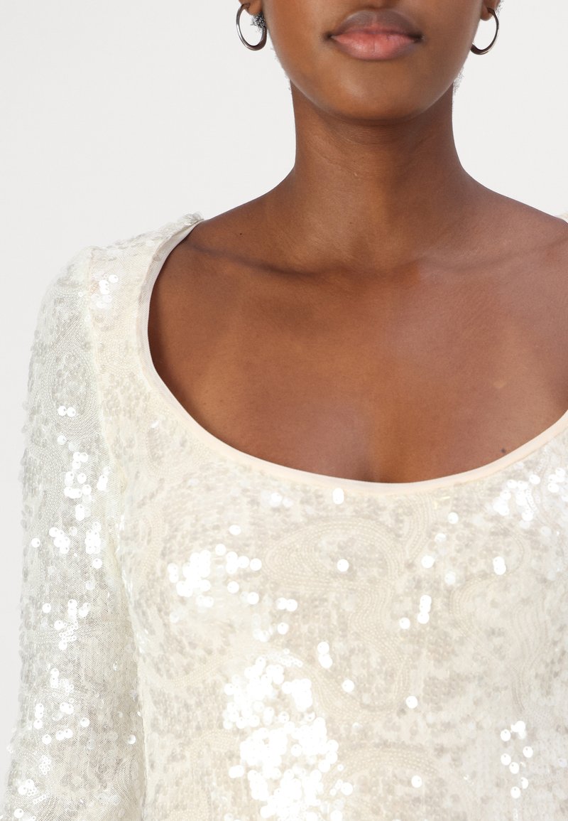 Top z cekinami o szerokim dekolcie, z teksturowym wzorem. Materiał jest kremowy, ozdobiony błyszczącymi, odbijającymi światło cekinami.